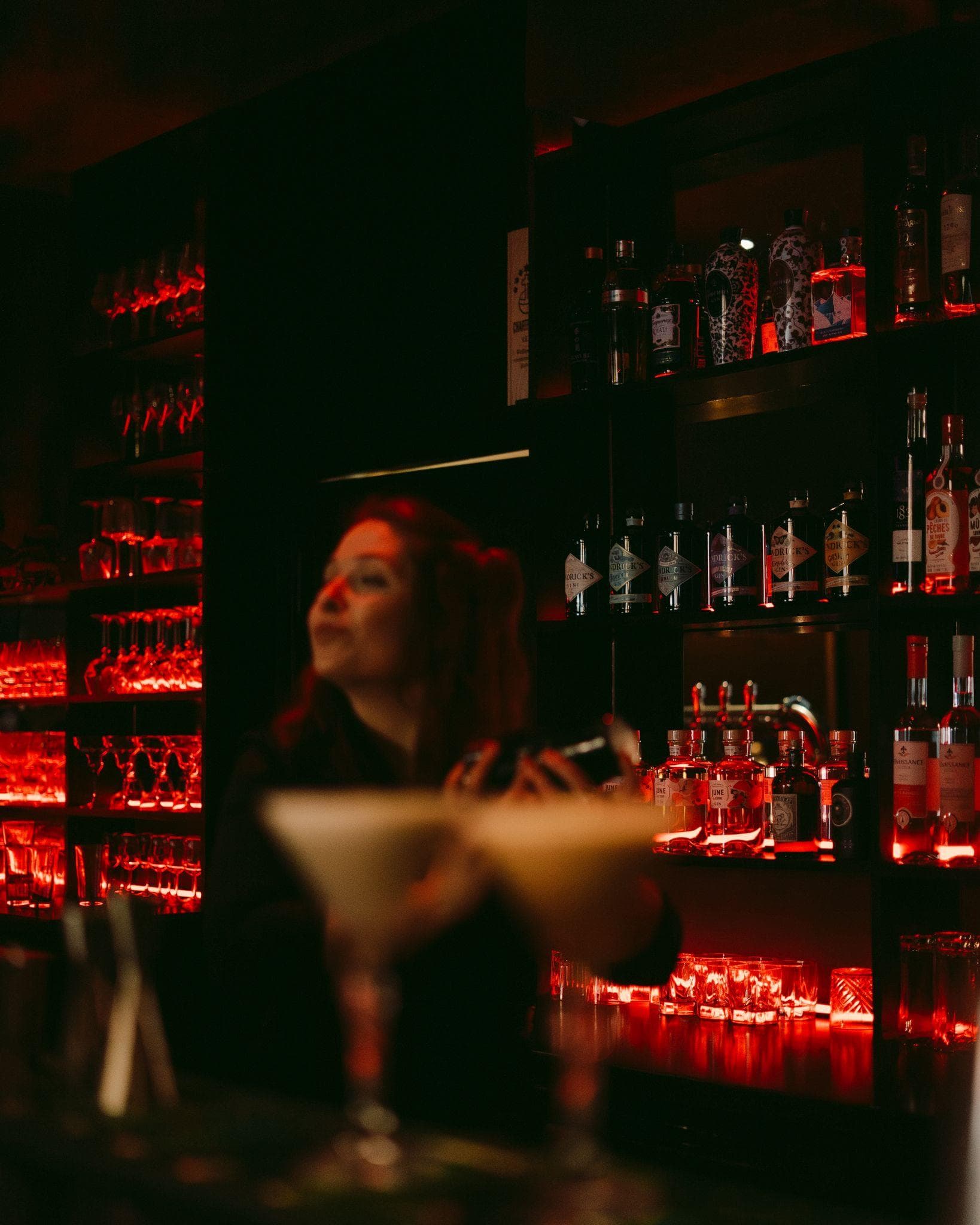 Barmaid derrière le bar du Bluebird, étagères de bouteilles baignées de lumière rouge